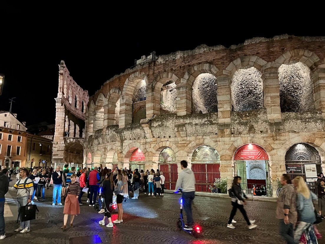 The illuminated Roman amphitheater at night, surrounded by visitors, with its arches glowing under warm lights.