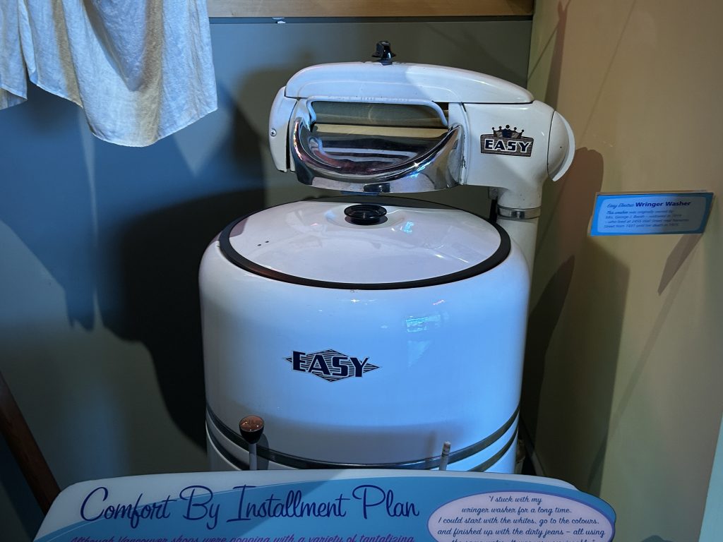 An antique "Easy" brand wringer washing machine displayed with a sign explaining its functionality. The machine is white with metallic accents and represents household innovations from the early 20th century.