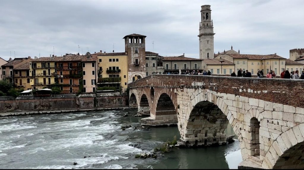 A historic Roman stone bridge crossing the Adige River, with flowing water beneath and Verona’s bell towers and colorful buildings in the background.