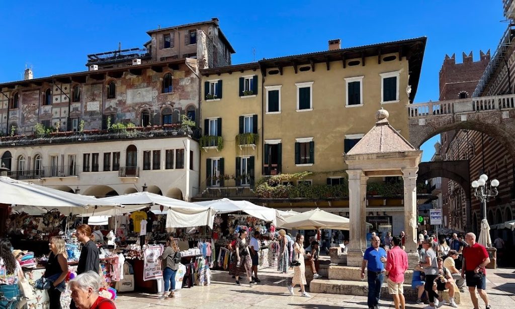 A lively outdoor market in Piazza delle Erbe, Verona, Italy, with white canopy tents filled with clothing, souvenirs, and accessories. People are strolling, browsing stalls, and chatting, while historic buildings with intricate frescoes and warm-toned facades create a picturesque backdrop. The Capitello, a small stone structure, stands in the foreground, adding to the charming and historic ambiance of the square.
